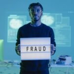 Man holding a 'FRAUD' sign in a tech setting, symbolizing cybersecurity threats.