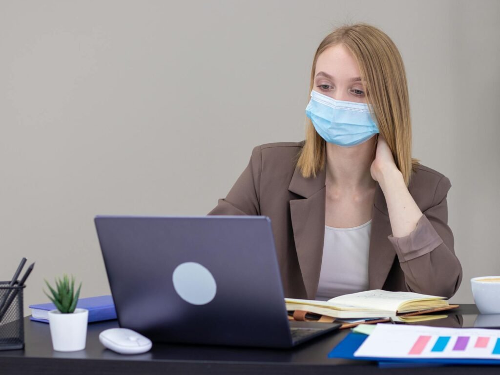 A professional woman wearing a mask works on a laptop in an office setting, ensuring safety.