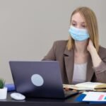 A professional woman wearing a mask works on a laptop in an office setting, ensuring safety.