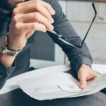Man in formal attire reviewing paperwork, holding glasses. Business setting.