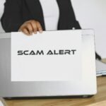 Business person holding a scam alert sign over a laptop, warning against online fraud.